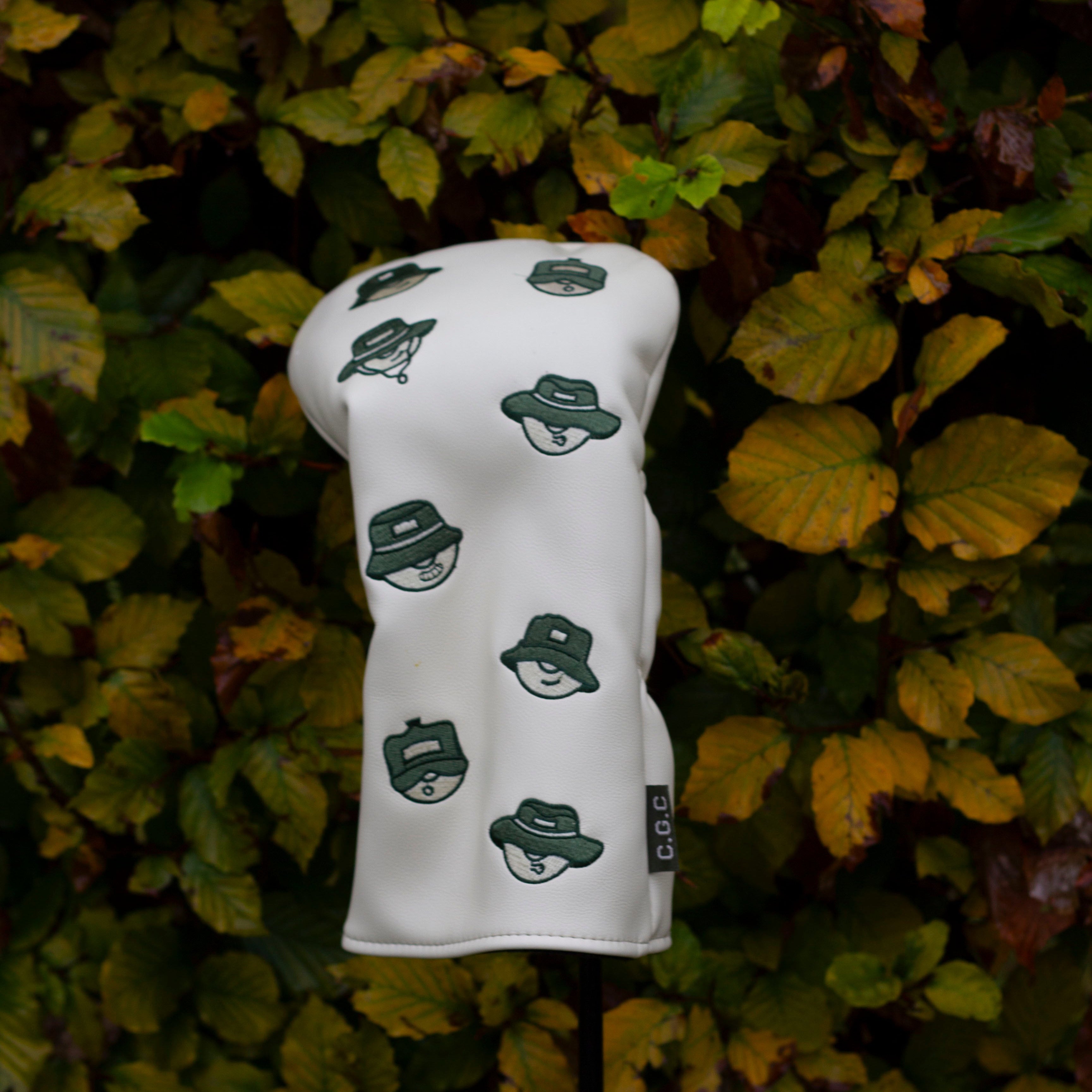 White golf club head cover with green hat pattern on a green leafy background